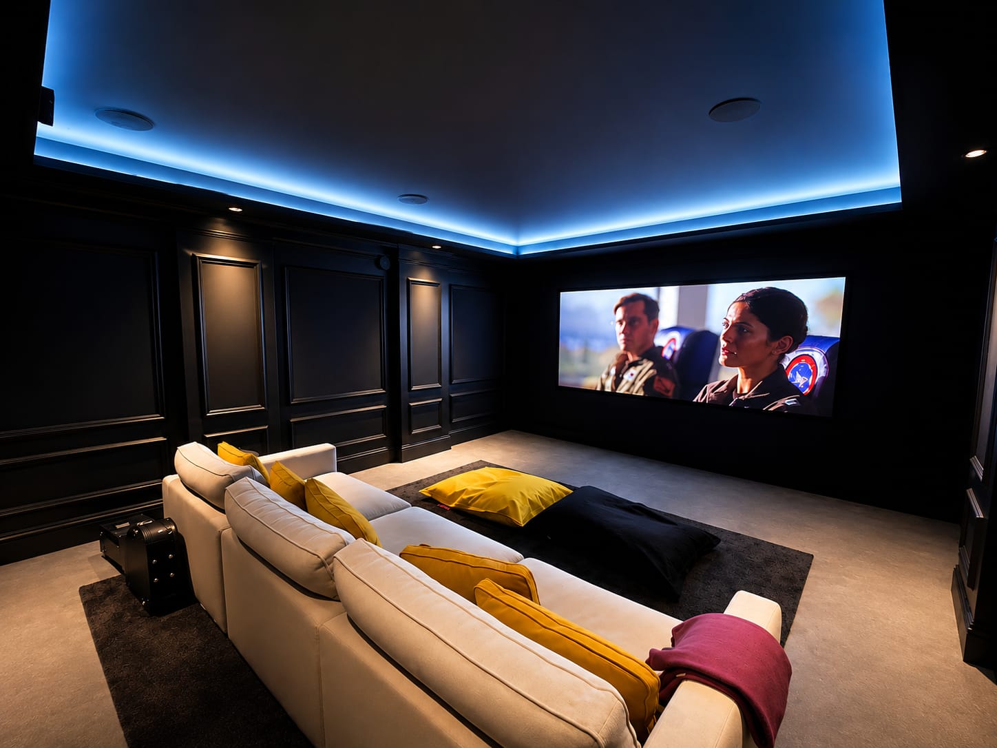 A dark home cinema with black panelled walls, a blue LED cove tracing the ceiling and a cream corner sofa facing a wide screen