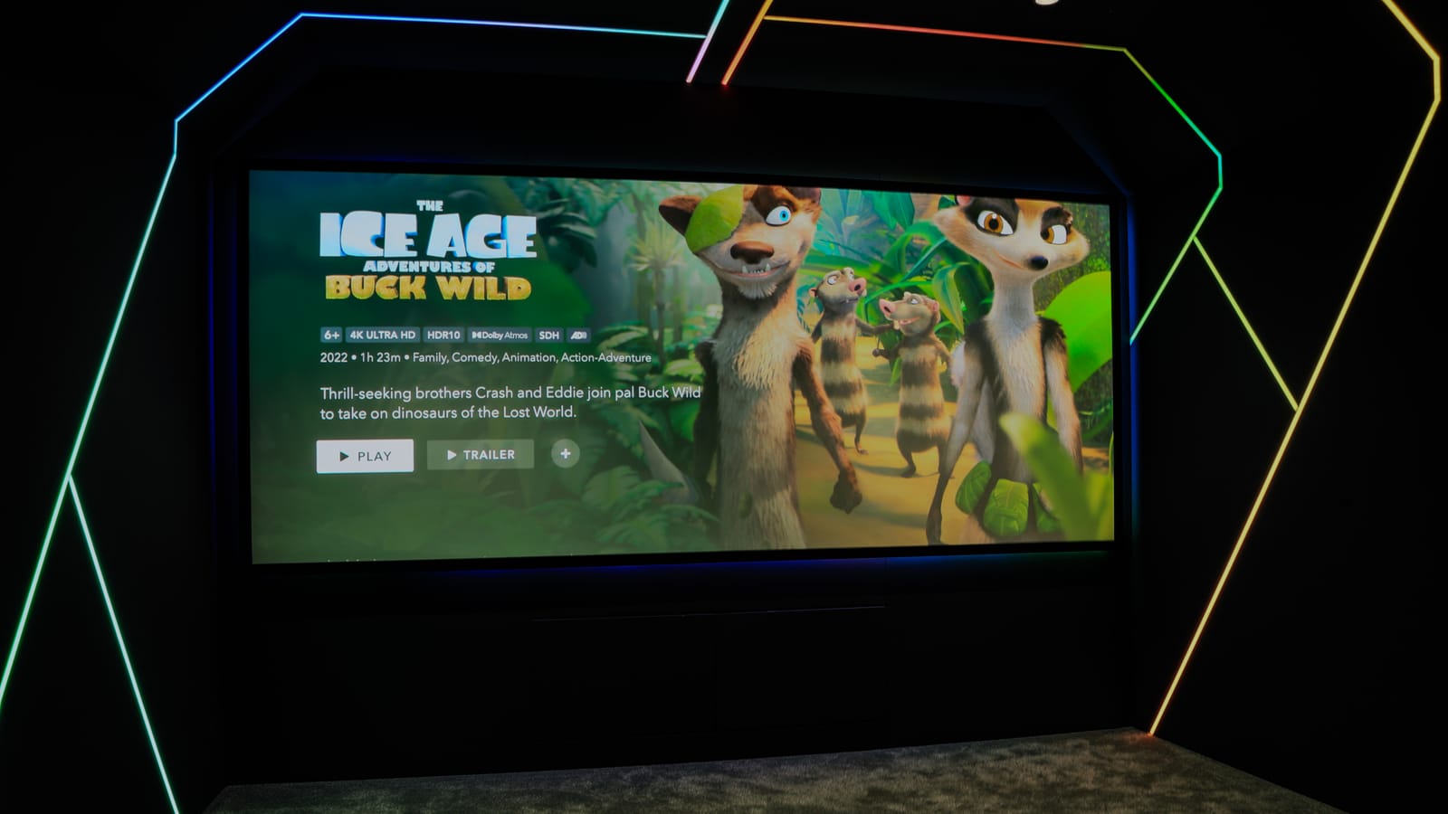 Entry-level cinema room with a wall-mounted screen framed by multi-colour LED strips, showing a film selection menu