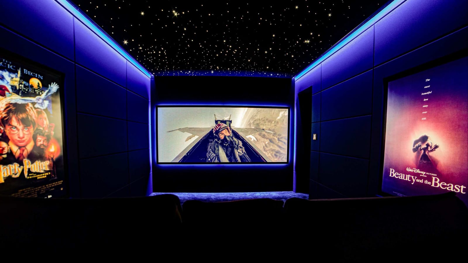 Mid-range home cinema: a dedicated cinema room viewed from the back row, with a fibre-optic starlight ceiling, blue cove lighting, framed film posters and a film playing