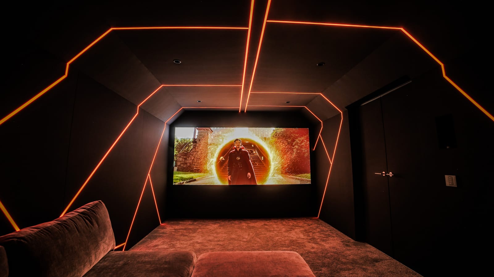 Dark home cinema with an illuminated orange geometric light feature tracing the walls and ceiling, and a film playing on the screen