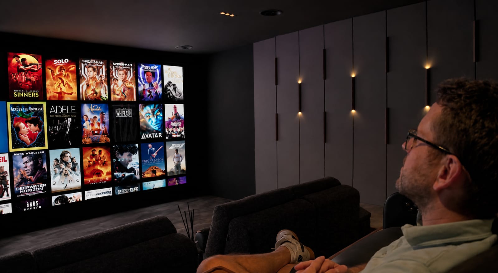 Home cinema installation by Acoustic Pixel: a man browsing a wall of cover art on the screen, lit by recessed wall lights