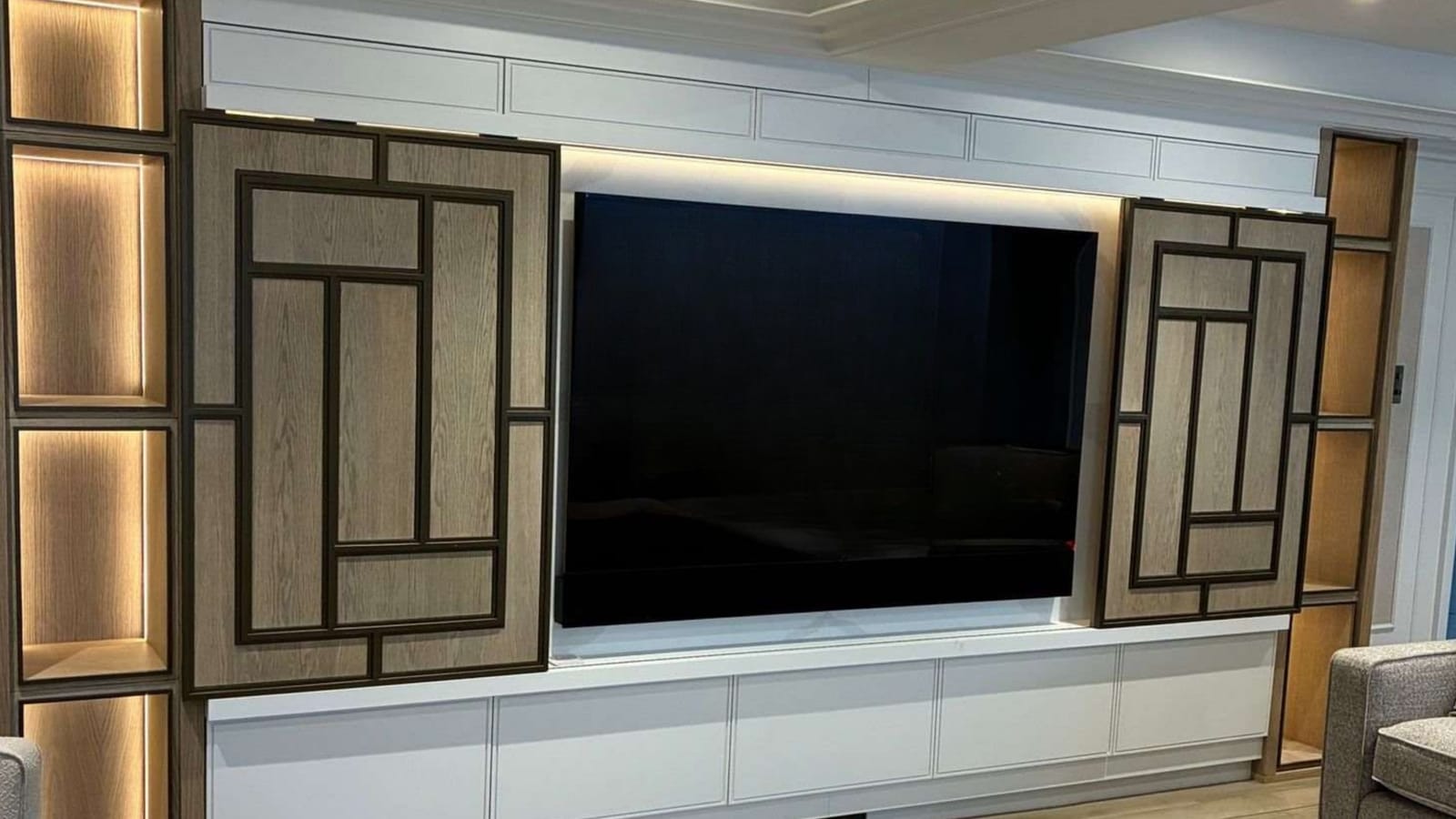 Finished media wall with a large flush-mounted television between oak fretwork panels, backlit display niches and a run of white base cabinets