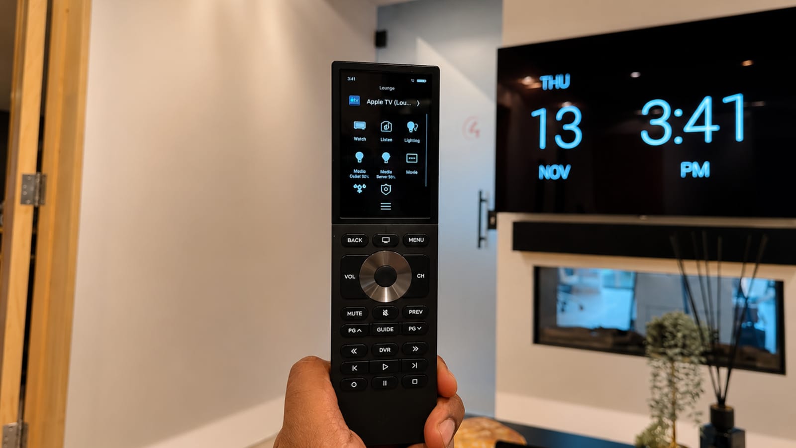 Hand holding a smart home remote whose touchscreen lists the room's sources and lighting, with a wall-mounted display showing the time behind